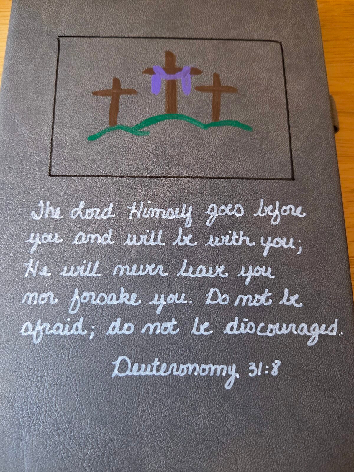 A hand-painted grey journal with three crosses and Deuteronomy 31:8.